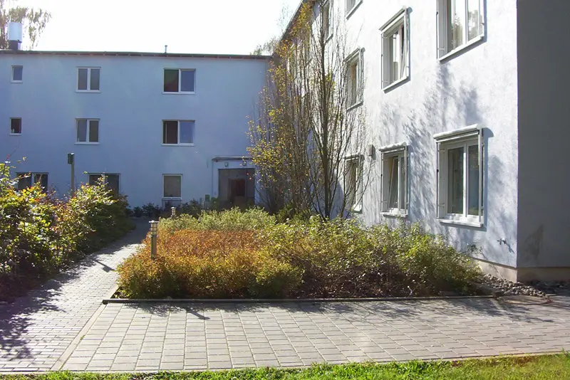 Innenhof mit gepflastertem Weg, Sträuchern und zwei weißen Gebäuden mit mehreren Fenstern.