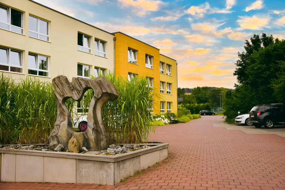 Skulptur aus verwittertem Holz in einem rechteckigen Pflanzbeet vor einem gelb-weißen mehrstöckigen Gebäude mit mehreren Fenstern und geparkten Autos auf gepflastertem Hof.