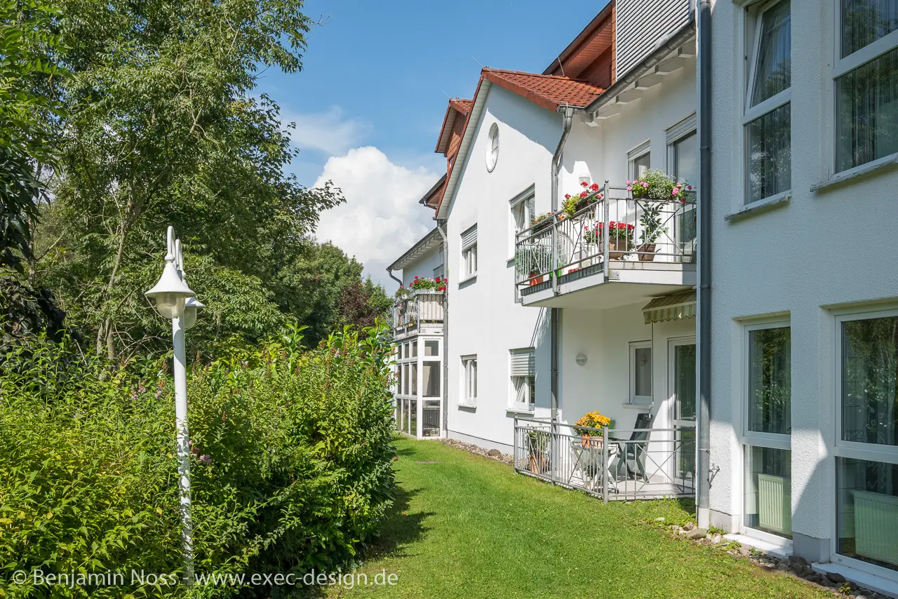 Außenansicht eines weißen Mehrfamilienhauses mit Balkonen und gepflegtem Garten bei Sonnenschein