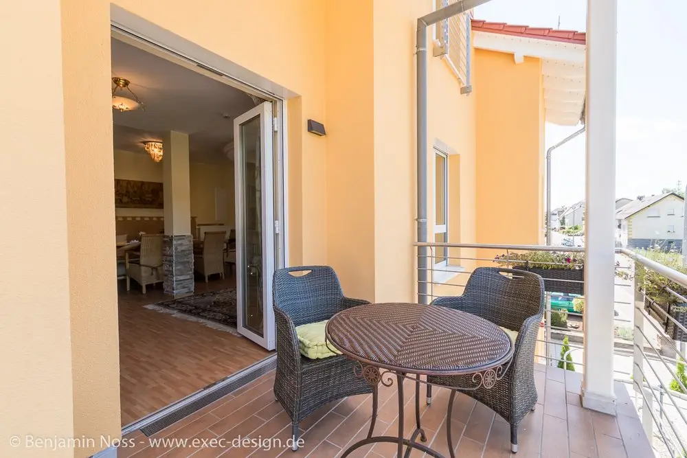 Balkon mit rundem Metalltisch und zwei geflochtenen Stühlen vor offenem Wohnzimmertür mit Blick ins Innere