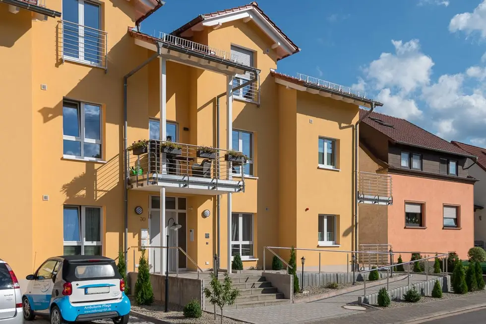 Mehrstöckiges Wohnhaus mit gelber Fassade, Balkonen und einer Rampe am Eingang bei sonnigem Wetter