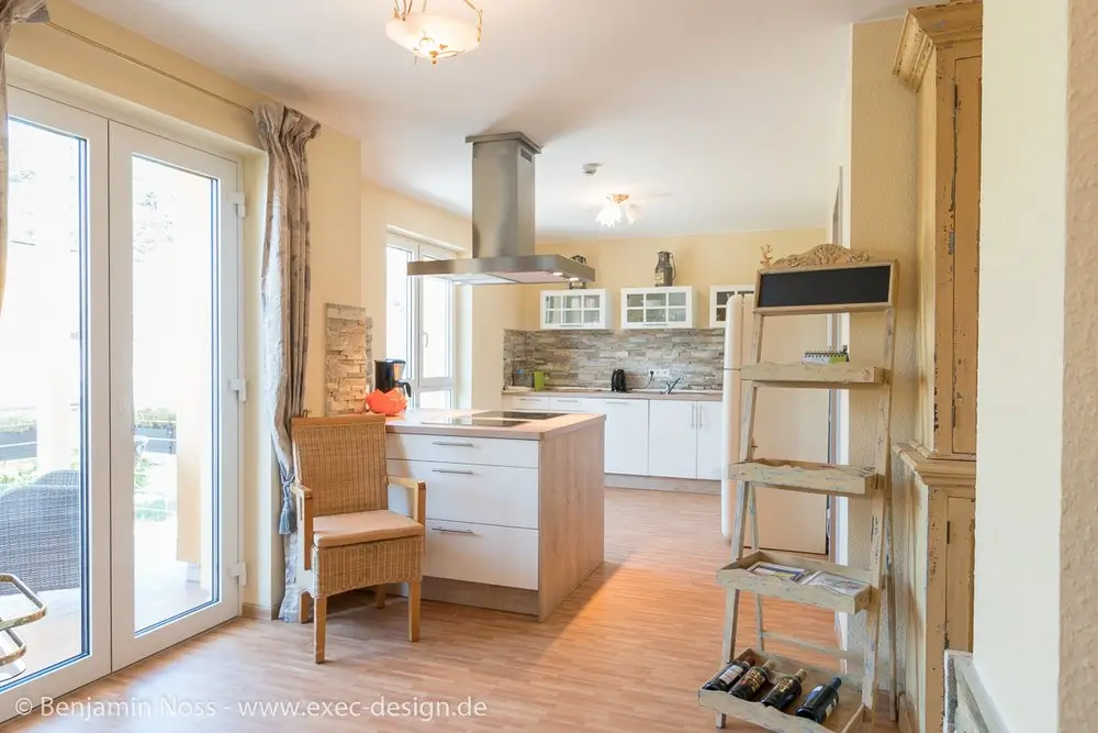 Helle moderne Küche mit Kochinsel, Korbstuhl, Holzregal mit Weinflaschen und großen Fenstertüren