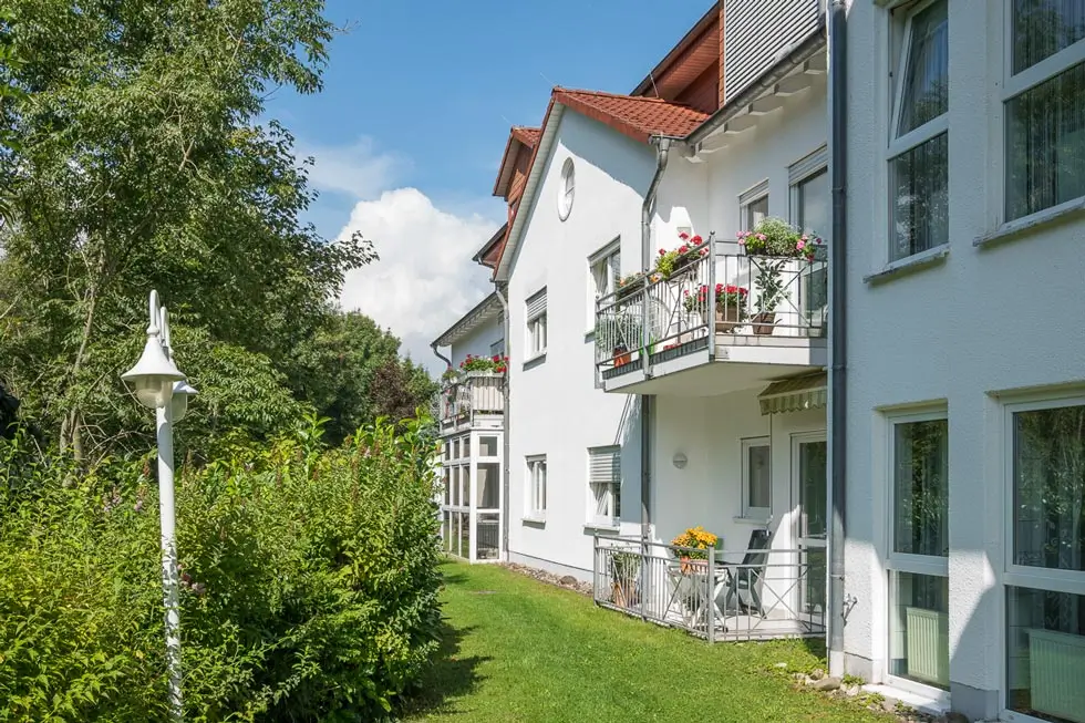 Weiße Wohnhäuser mit Balkonen und Blumen neben einem gepflegten Rasen und Büschen unter blauem Himmel