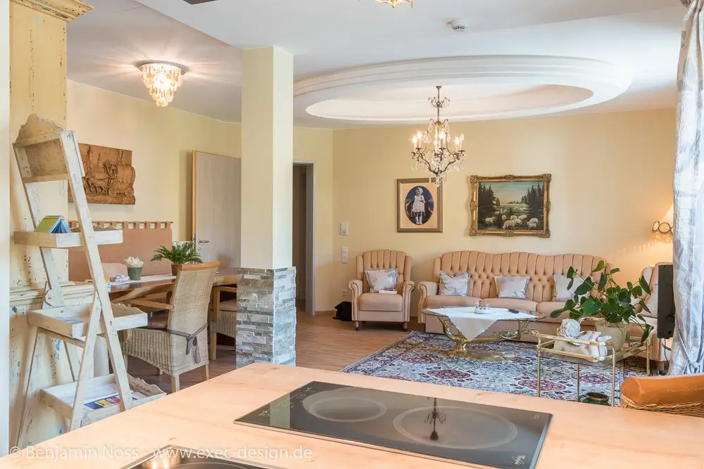 Offene Wohnküche mit Kochfeld im Vordergrund, Essbereich mit Holztisch und Korbstühlen sowie Wohnzimmer mit Sofa, Sesseln, Teppich und Kronleuchtern