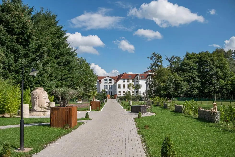 Breiter gepflasterter Weg in einem Park mit Bänken, Bäumen und einem weißen Gebäude im Hintergrund unter blauem Himmel mit Wolken