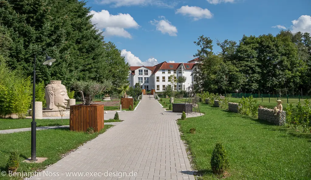 Breiter gepflasterter Weg in einem Park mit Bänken, Bäumen und einem weißen Gebäude im Hintergrund unter blauem Himmel mit Wolken